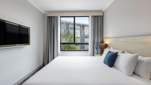 Habitación de hotel con una cama grande, un televisor de pantalla plana montado en la pared, una mesita de noche con lámpara y una ventana con cortinas.