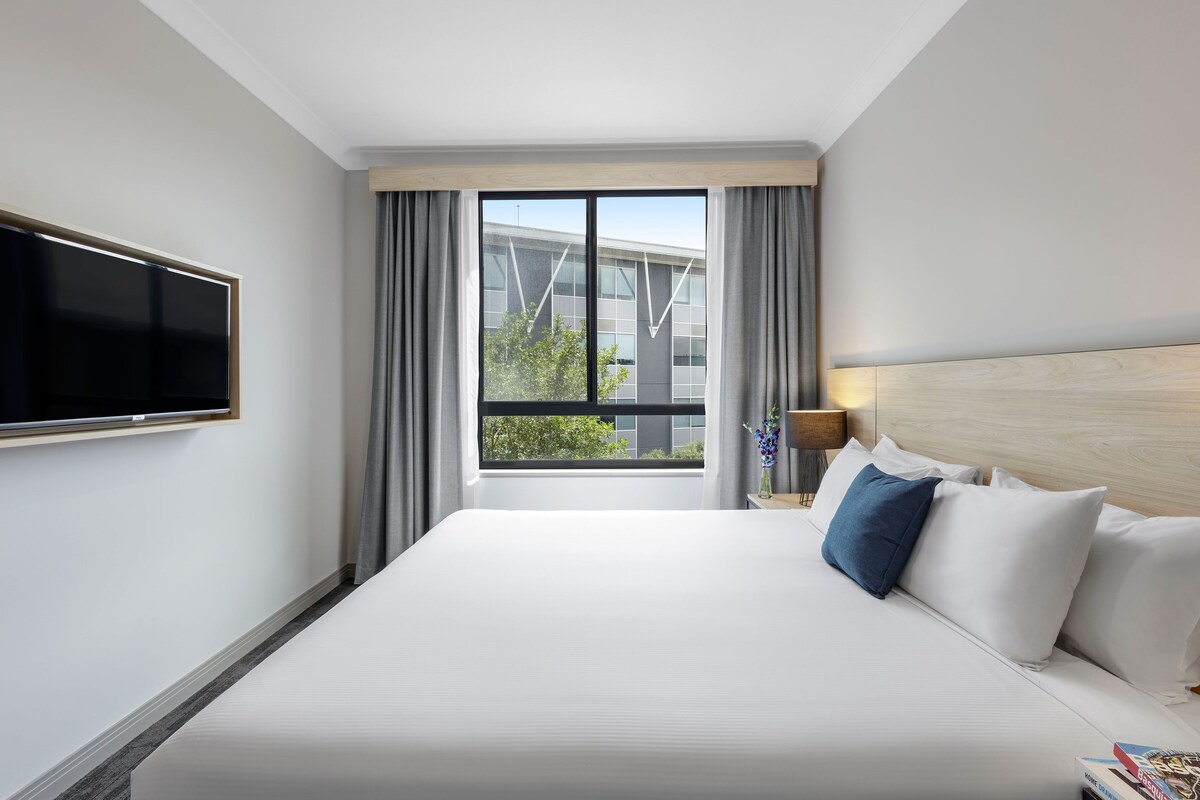 A hotel room with a large bed, a flat-screen TV mounted on the wall, a nightstand with a lamp, and a window with curtains.