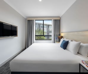 Habitación de hotel con una cama grande, un televisor de pantalla plana montado en la pared, una mesita de noche con lámpara y una ventana con cortinas.