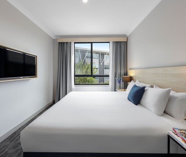 A hotel room with a large bed, a flat-screen TV mounted on the wall, a nightstand with a lamp, and a window with curtains.