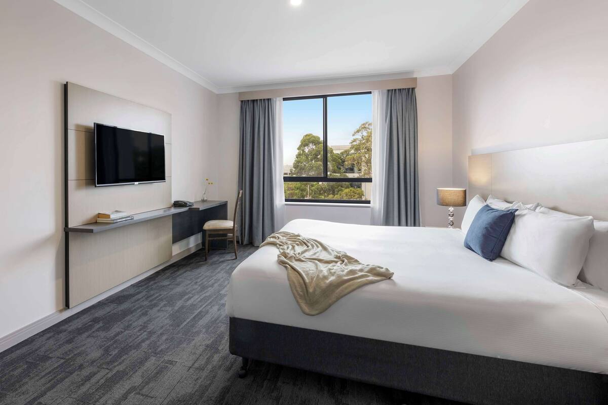 A hotel room with a bed, a TV mounted on the wall, a desk, and a window with curtains.