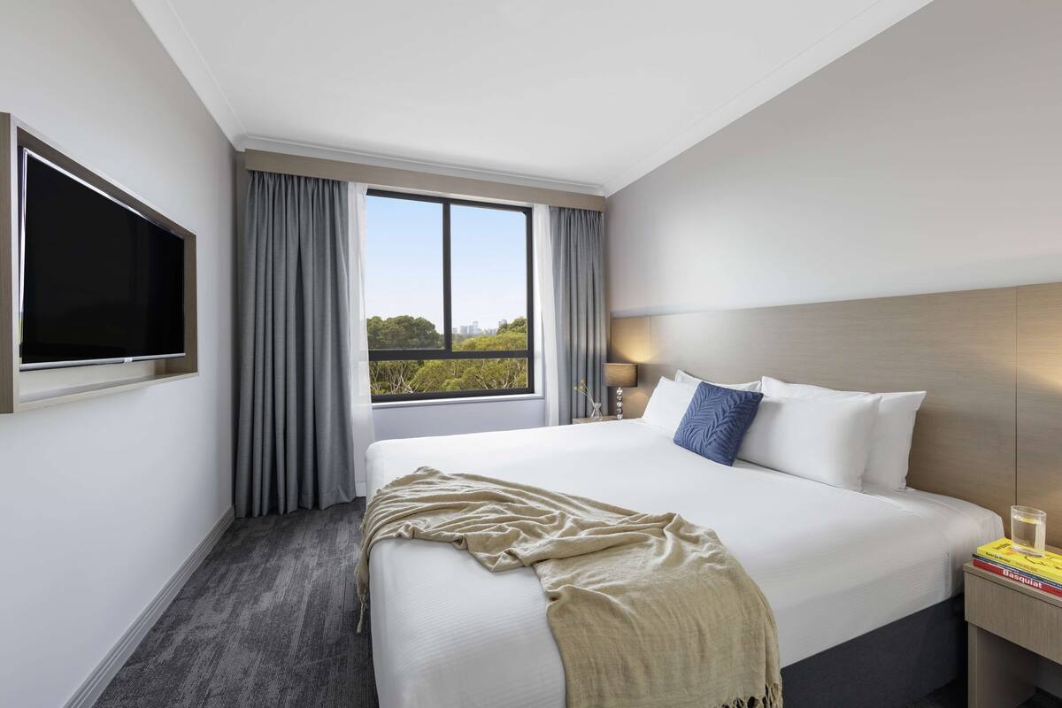A hotel room with a large bed, a TV mounted on the wall, a window with curtains, and a bedside table with a lamp and books.