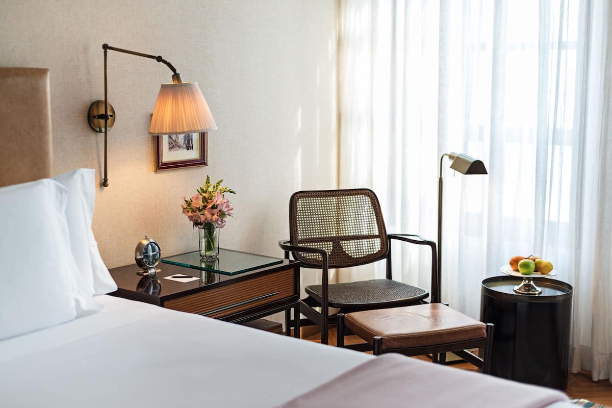 Una habitación de hotel con una cama, una silla, una mesita con una lámpara, un cuadro en la pared y un florero con flores.
