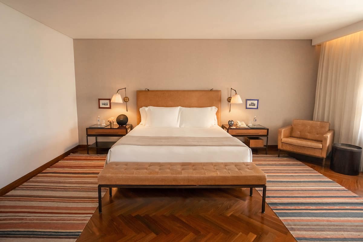 Una habitación de hotel con una cama grande, dos mesitas de noche con lámparas, una silla y suelo de madera.