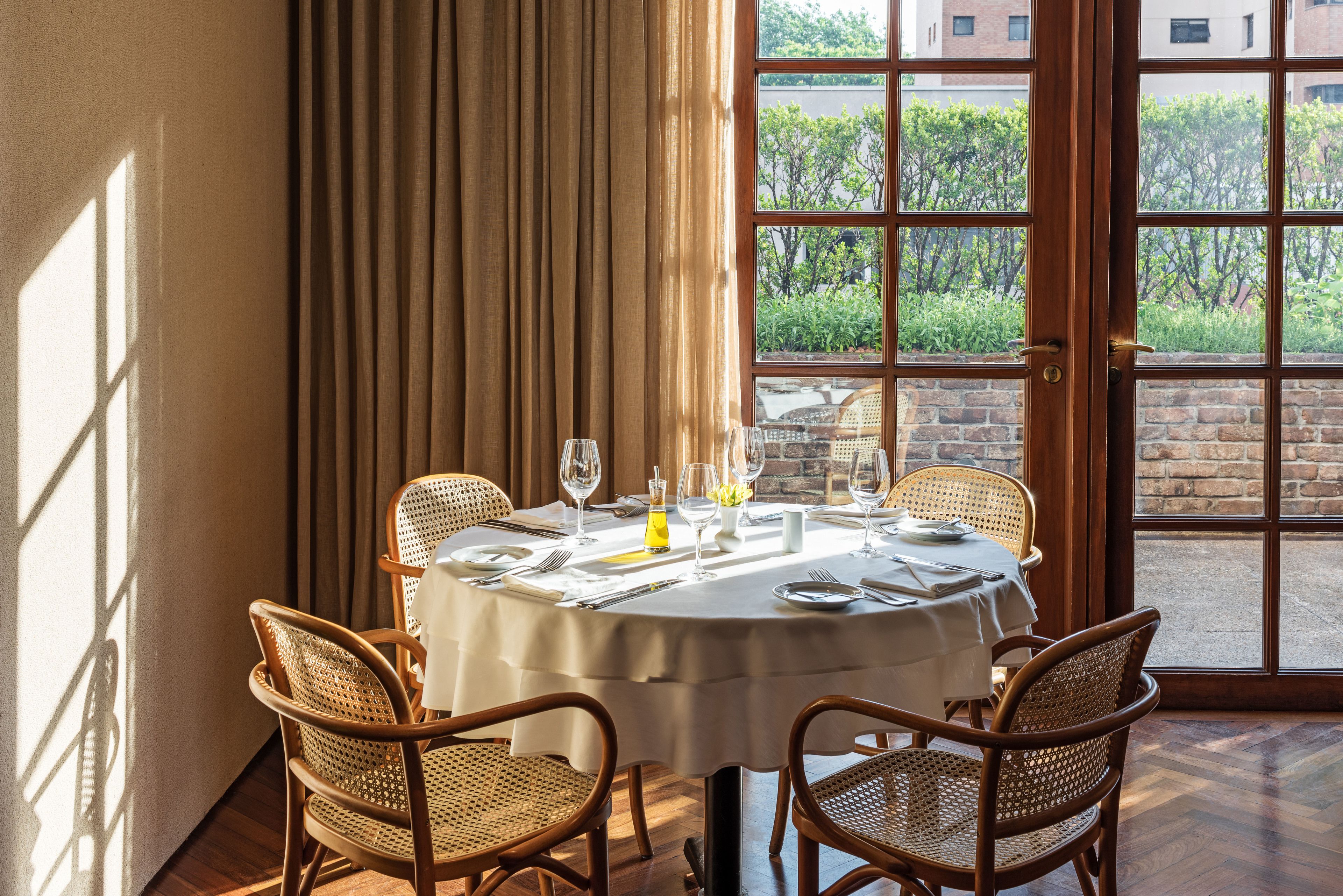 A round dining table set for four with wicker chairs, a white tablecloth, and a view of greenery through a large window.