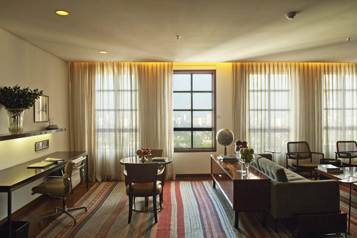 Habitación de hotel con escritorio, silla, sofá y vistas a la ciudad a través de amplios ventanales.