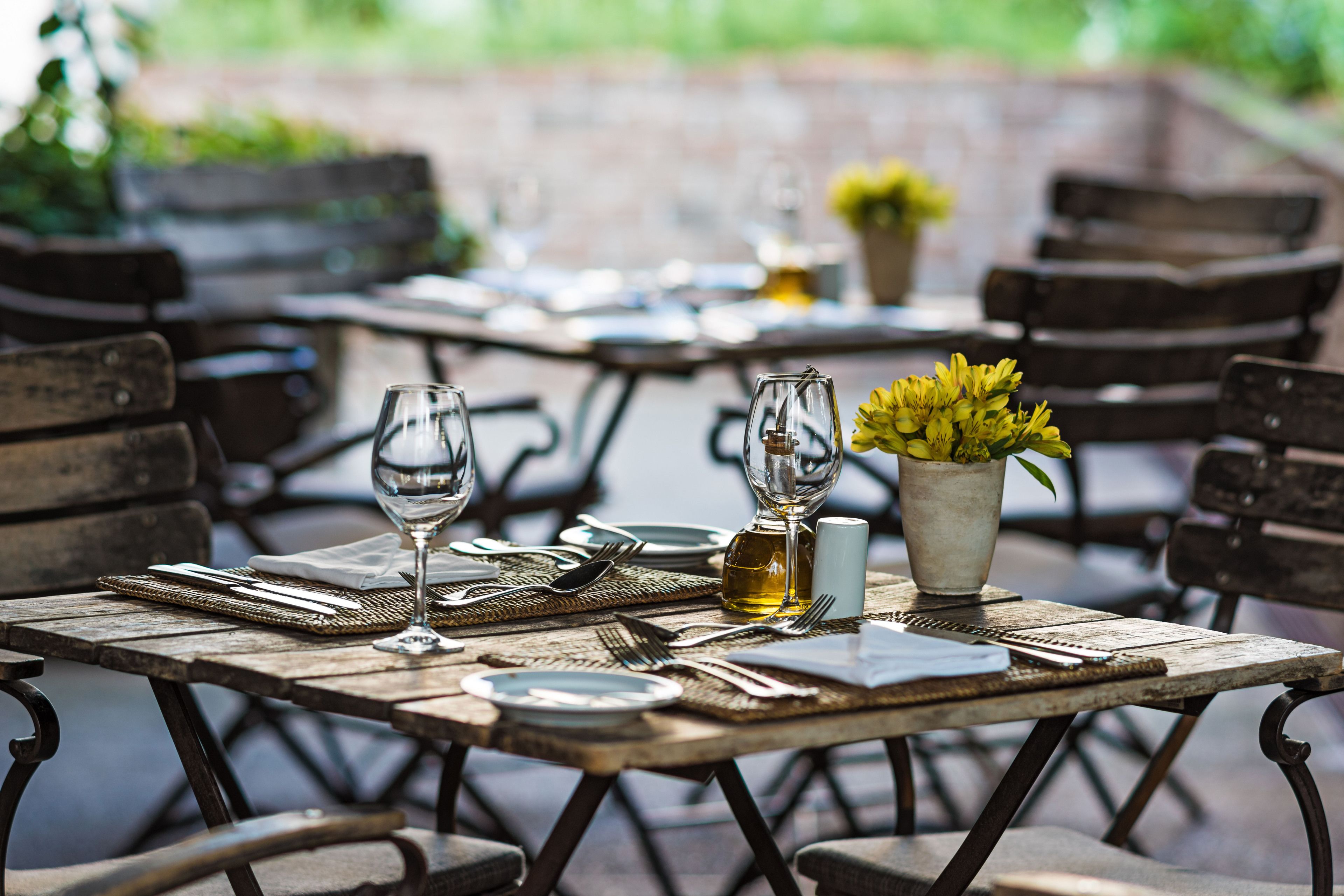 An outdoor dining area with wooden tables and chairs, equipped with wine glasses, cutlery, and a vase with yellow flowers.