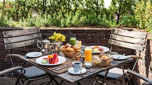 Un área de comedor al aire libre con pared de ladrillo, bancos de madera y una mesa dispuesta con productos para el desayuno, como cruasanes, fruta y una bebida.