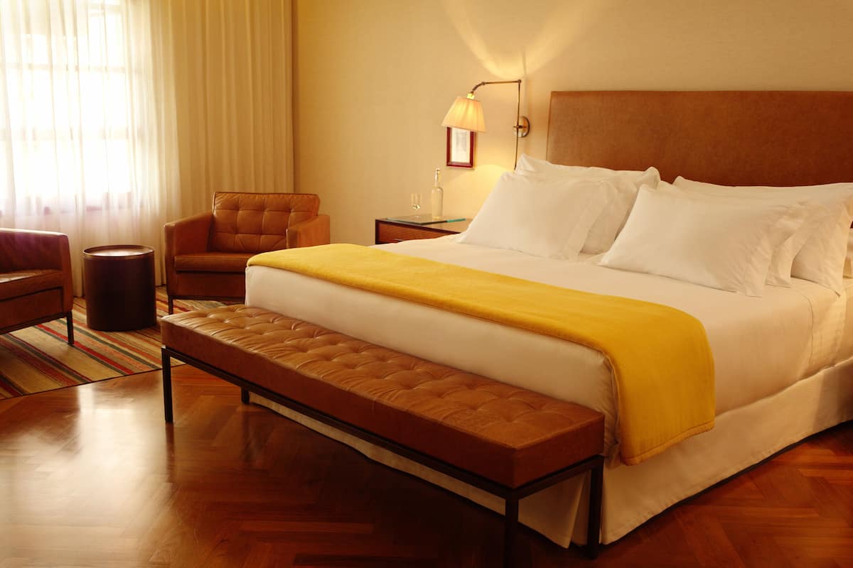 Una habitación de hotel con una cama grande, un banco, una silla, una mesita de noche y una lámpara.