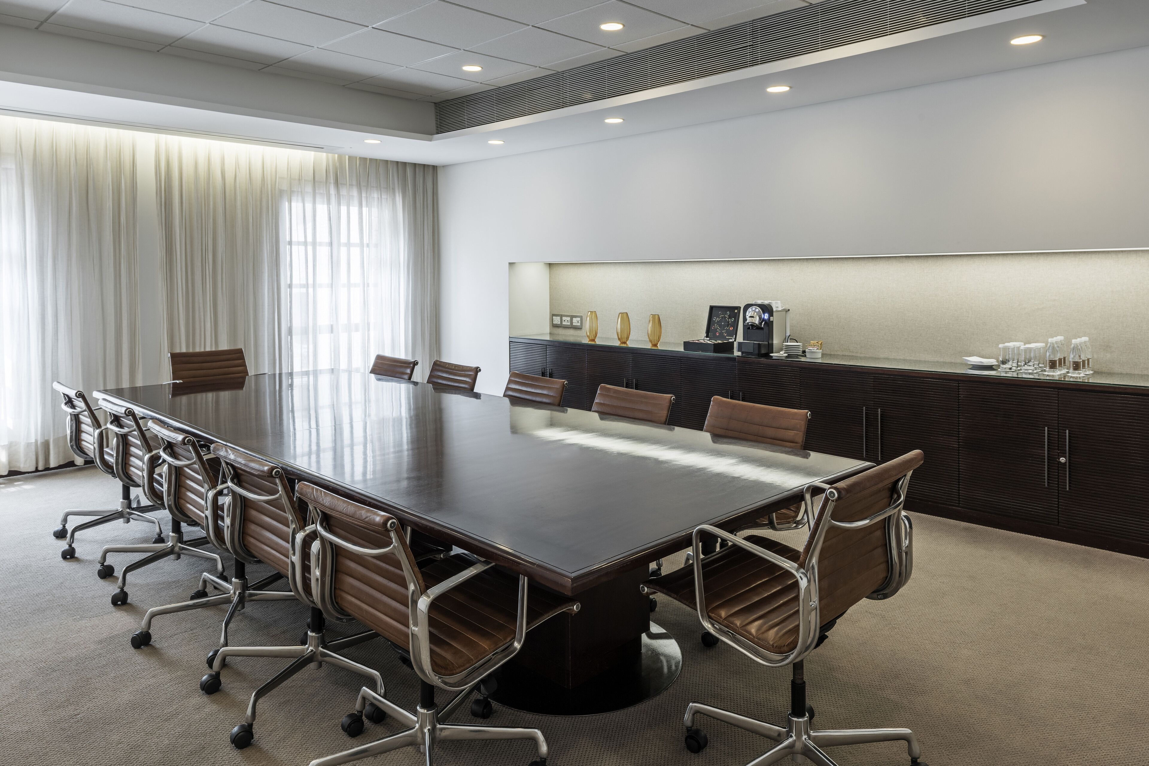 A conference room with a long table, chairs, a dark wooden cabinet, and a window with curtains.