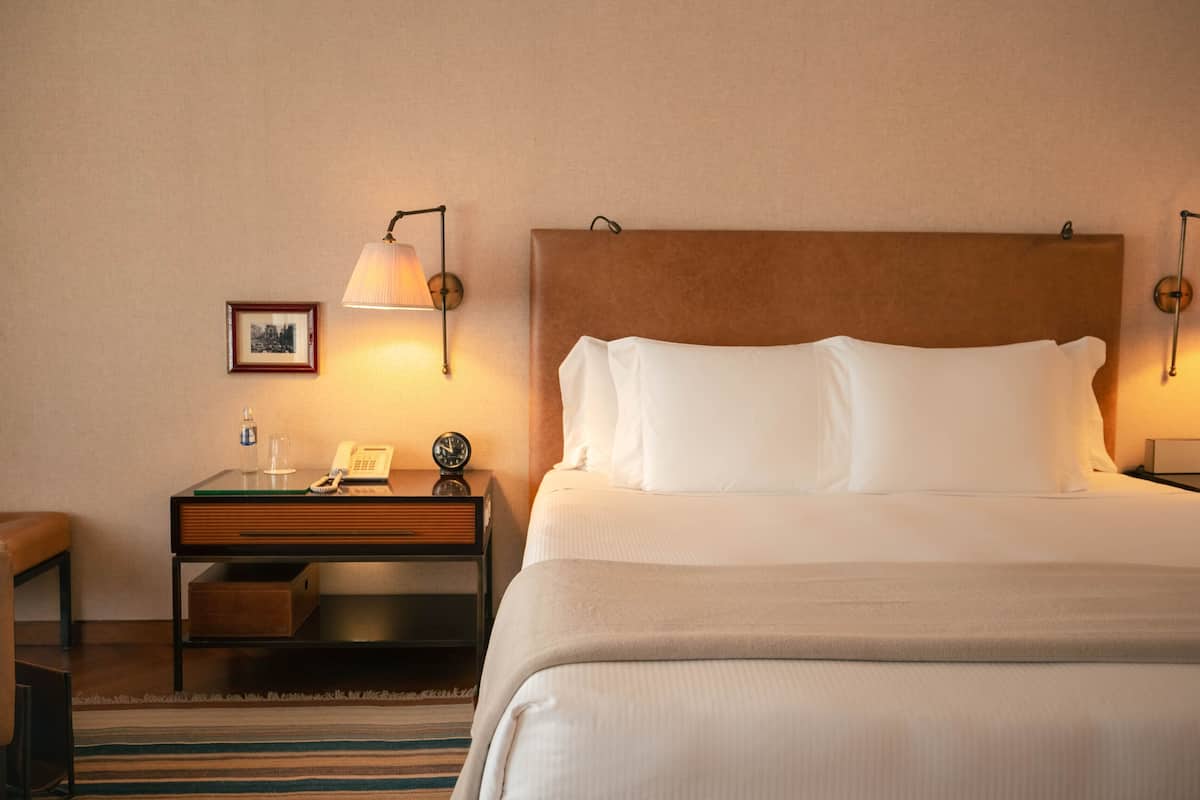Habitación de hotel con una cama grande, una mesita de noche con lámpara, un cuadro en la pared y una pequeña mesita con teléfono y reloj.