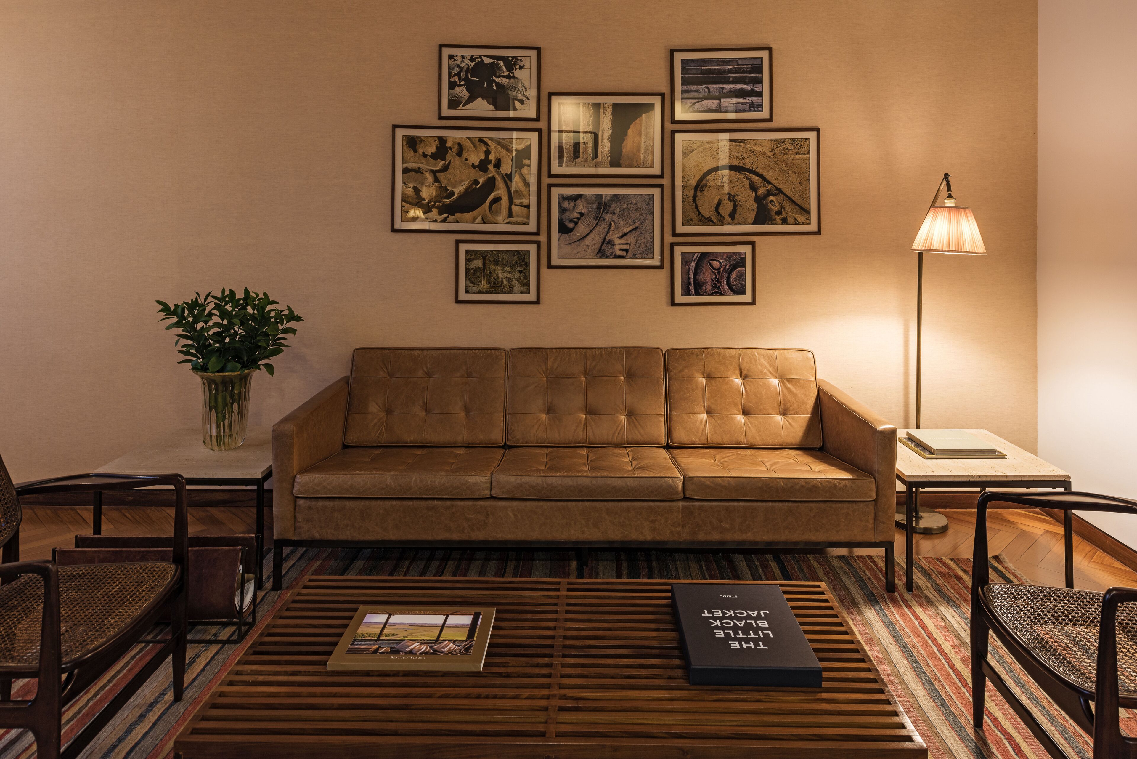Una sala de estar con un sofá de cuero marrón, una mesita de centro con revistas, una lámpara de pie y una pared decorada con marcos.