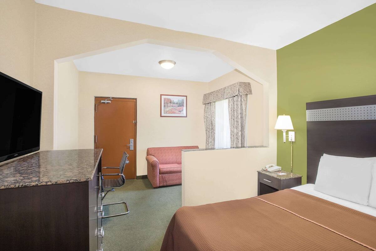 Ein Hotelzimmer mit einem Bett, einem Schreibtisch mit Stuhl, einem Fernseher, einem Fenster mit Vorhängen und einem Nachttisch mit Lampe.
