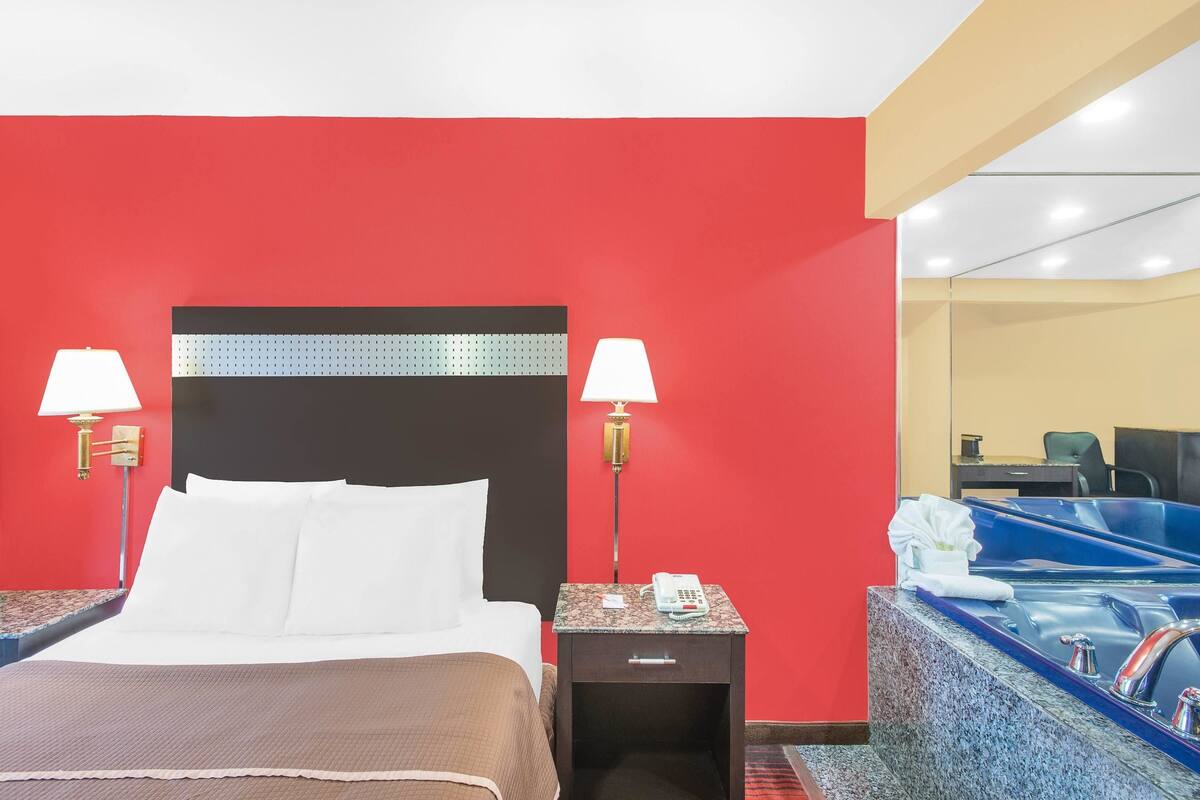 Ein Hotelzimmer mit roter Wand, einem Bett mit weißer Bettwäsche, zwei Nachttischlampen, einem Telefon, einer kleinen Schublade und einem Badezimmer mit einer blauen Badewanne und Toilette.