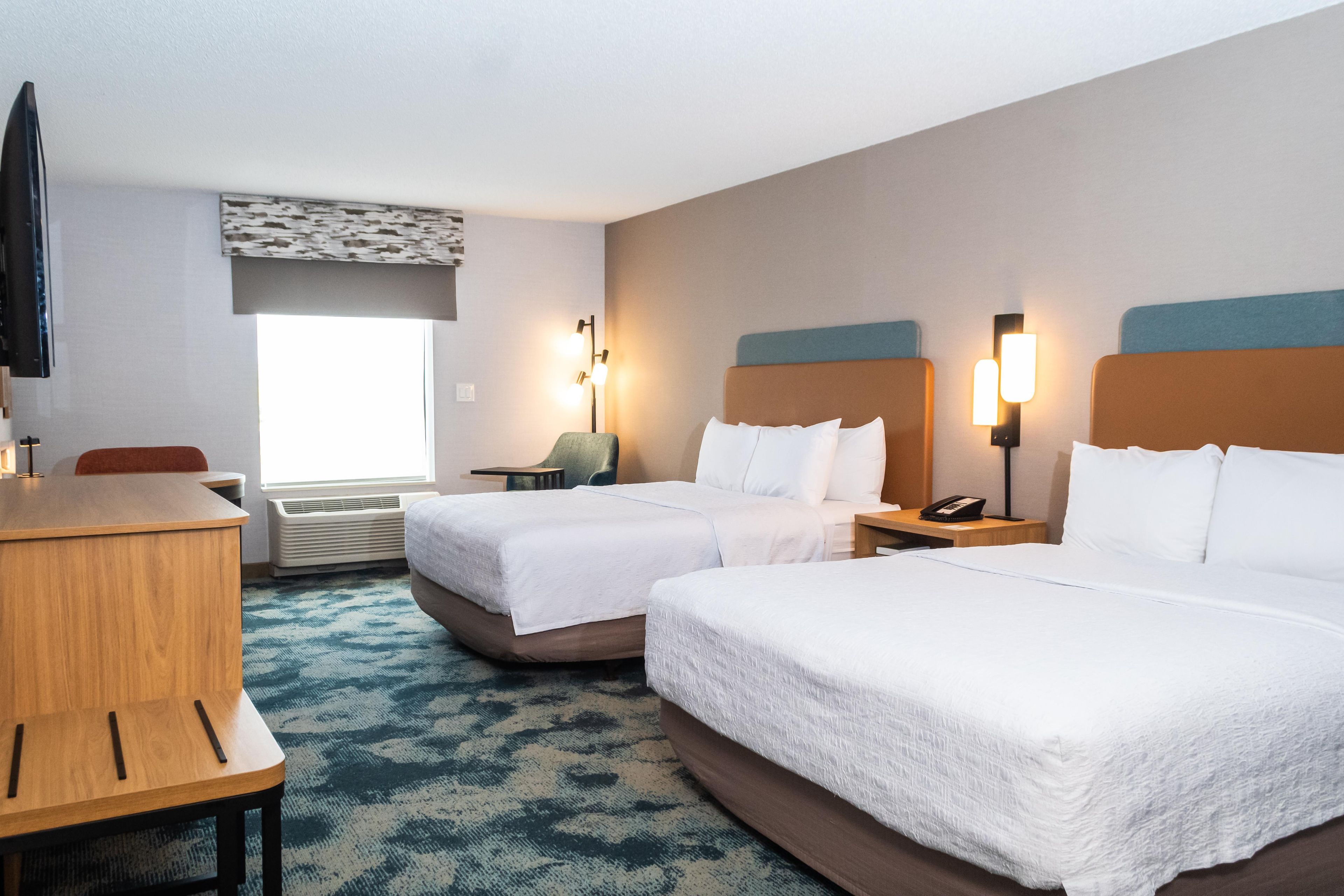 A hotel room with two beds, a desk, a chair, a TV, and a window with blinds.
