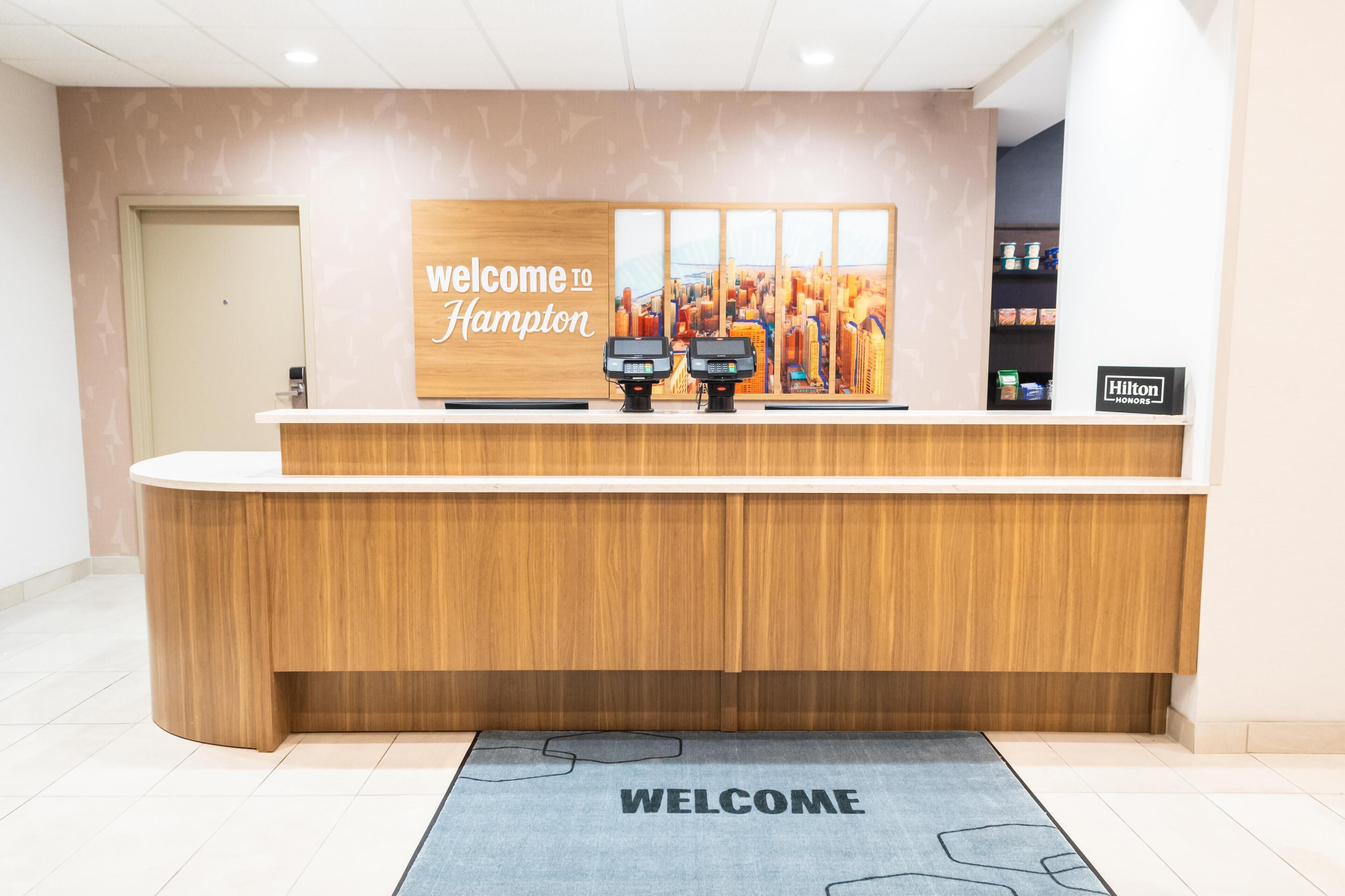Balcão de recepção de hotel com uma placa 'Welcome to Hampton', dois leitores de cartão e vista para a paisagem urbana.