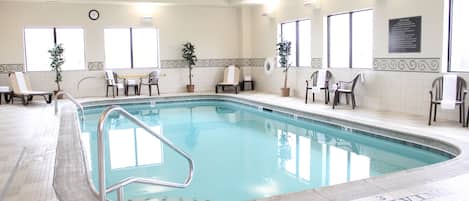 Una piscina interior con un cartel de prohibición de saltos, sillas, plantas en macetas y un reloj en la pared.