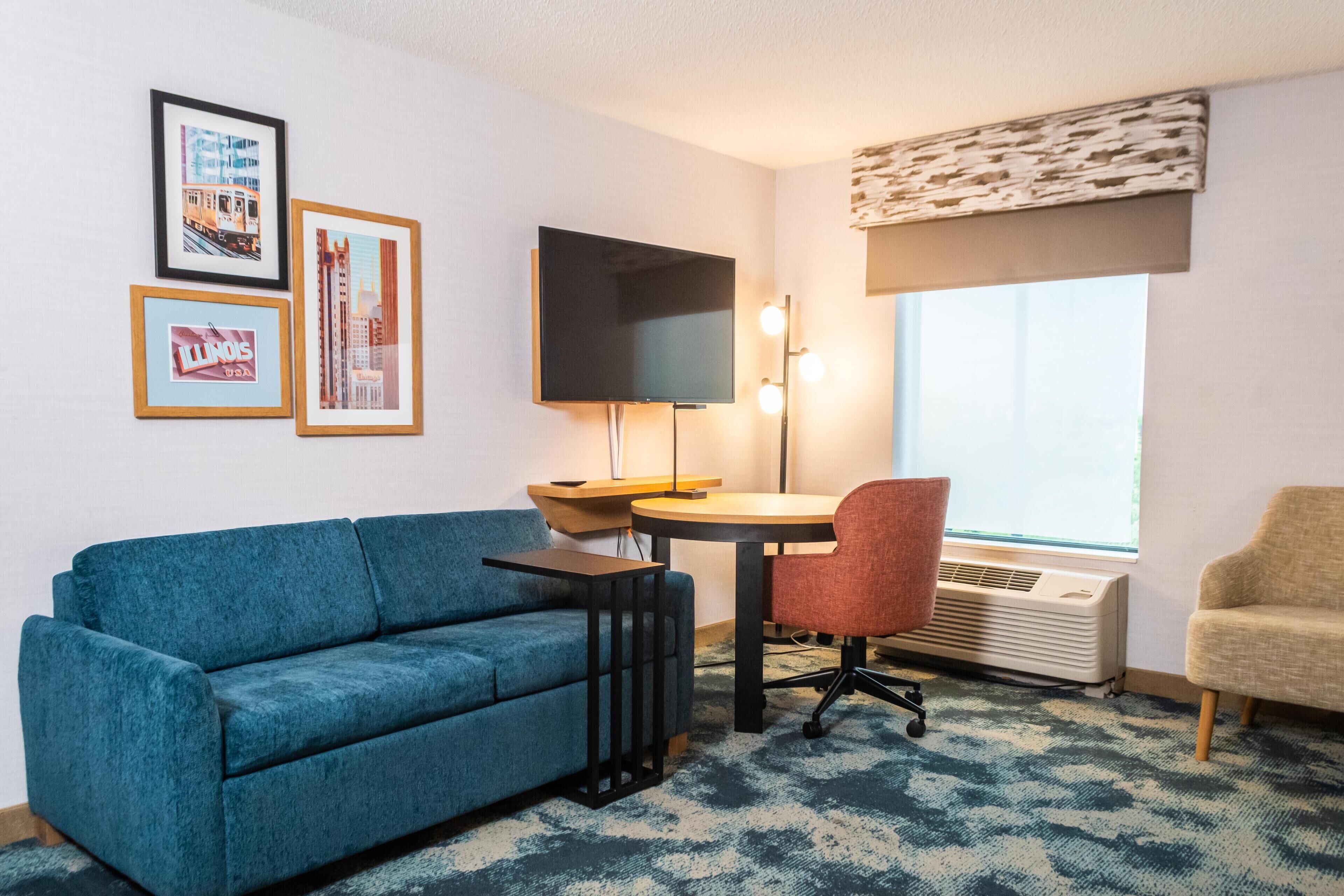 Una habitación con un sofá azul, una mesita redonda pequeña, una silla roja, un televisor montado en la pared y cuadros enmarcados.
