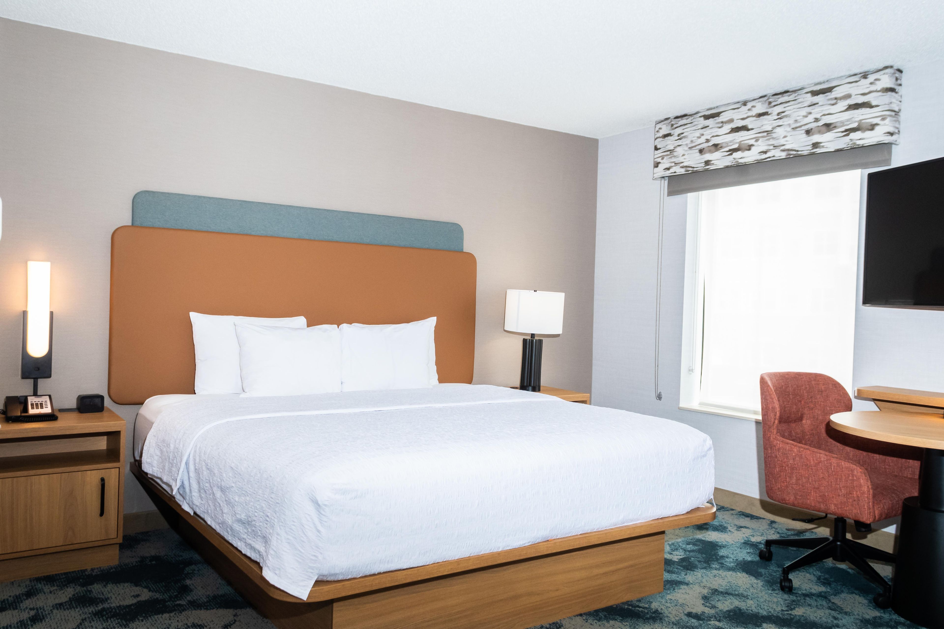 A hotel room with a bed, bedside tables, a desk, a chair, a lamp, and a TV.