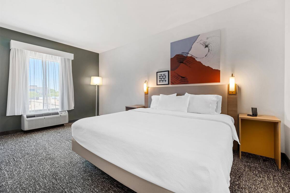 Hotellihuone, jossa on suuri sänky, ikkuna valkoisilla verhoilla, seinälle ripustettu taideteos ja kaksi yöpöytälamppua.