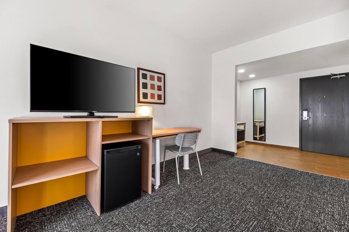 Moderni hotellihuone, jossa on taulutelevisio, työpöytä tuolilla, pieni jääkaappi ja matolla peitetty lattia.