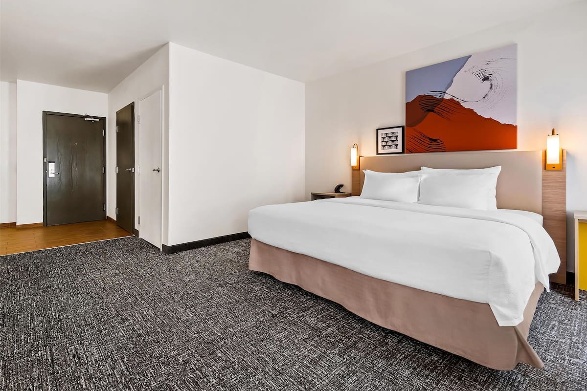 Hotellihuone, jossa on suuri sänky, seinällä taulu ja yöpöydällä lamppu.