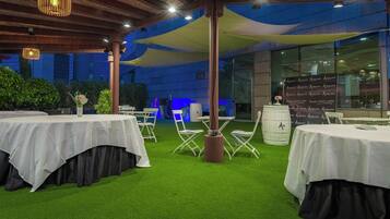 An outdoor event space with tables covered in white tablecloths, green artificial turf, and white folding chairs.