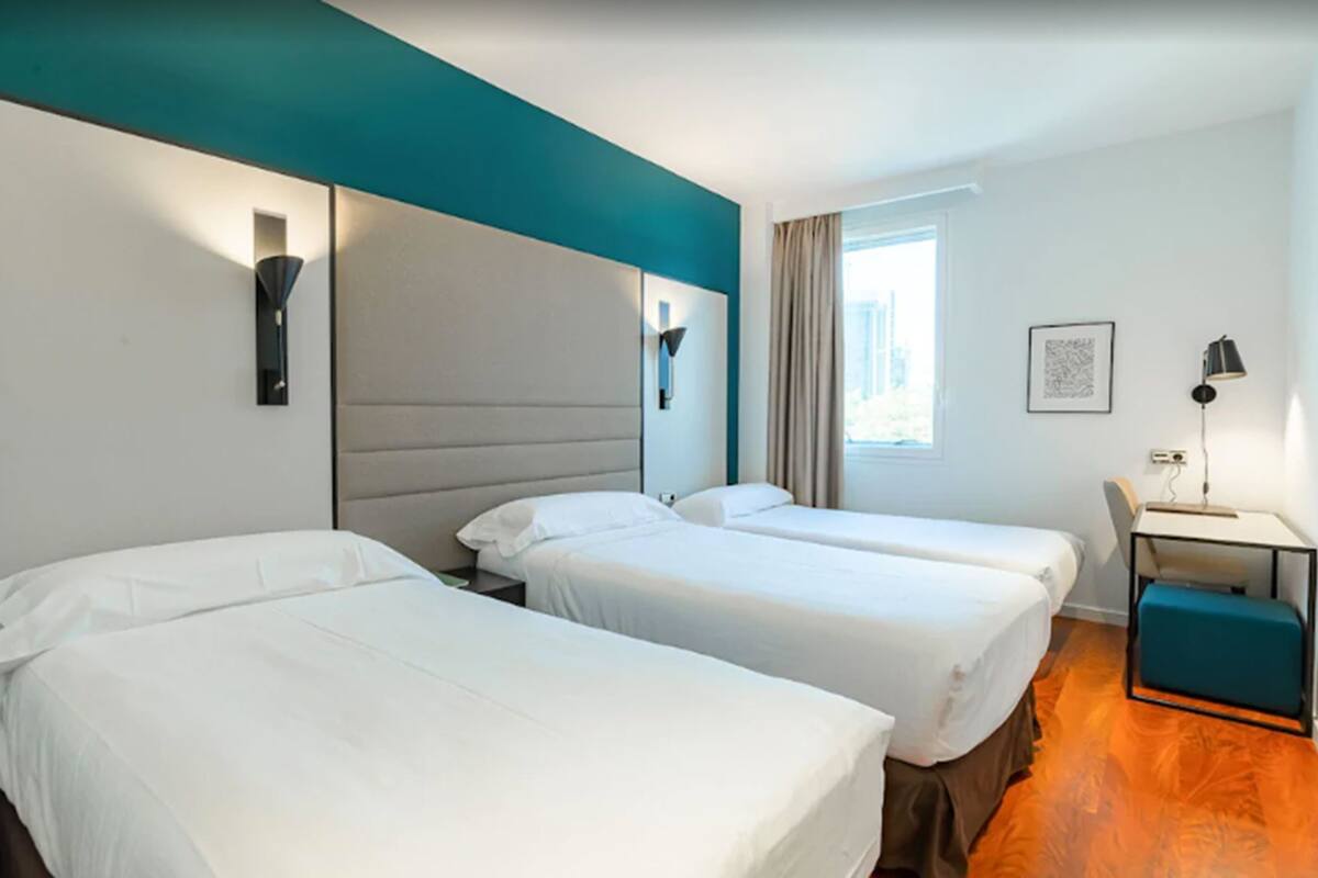 Une chambre d’hôtel avec deux lits simples, un bureau avec une chaise, une fenêtre avec des rideaux et une lampe fixée au mur.