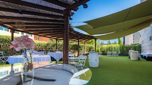 Een terras buiten met ronde tafels, witte parasols en een glazen tafel met een bloevas.