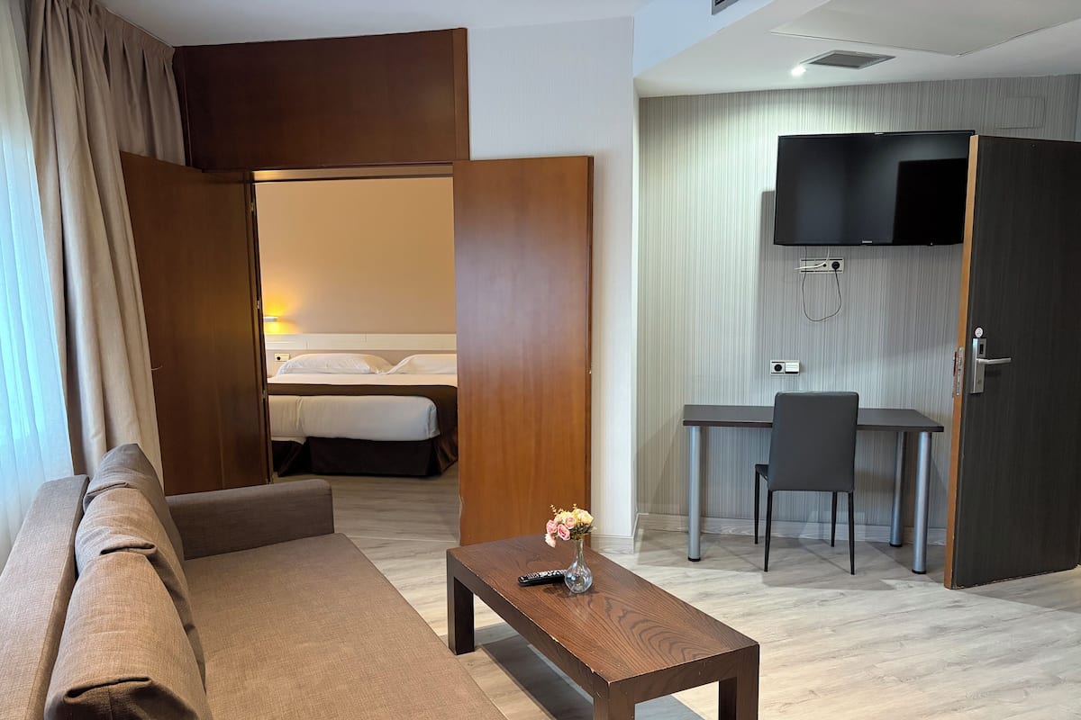 Une chambre d’hôtel avec un canapé, une petite table en bois sur laquelle est posé un vase, un lit avec du linge de maison blanc, un bureau avec une chaise, un téléviseur fixé au mur et une porte avec une poignée.