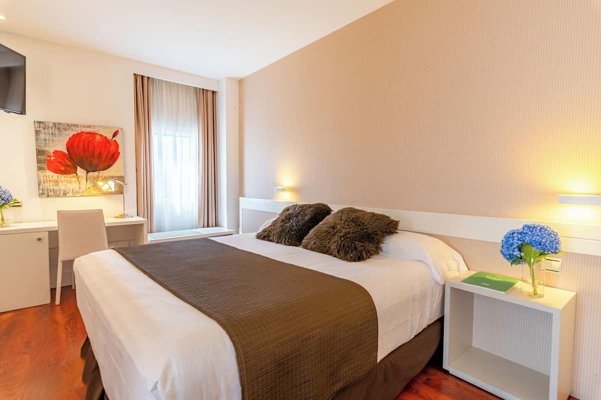 Une chambre d’hôtel avec un lit, un bureau, une chaise, une télévision et une fenêtre avec des rideaux.