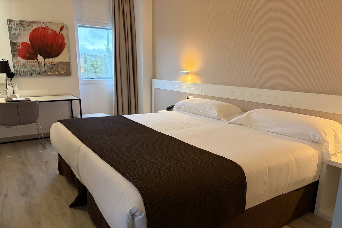 Une chambre d’hôtel avec un grand lit, un bureau, une chaise, un tableau et une fenêtre.