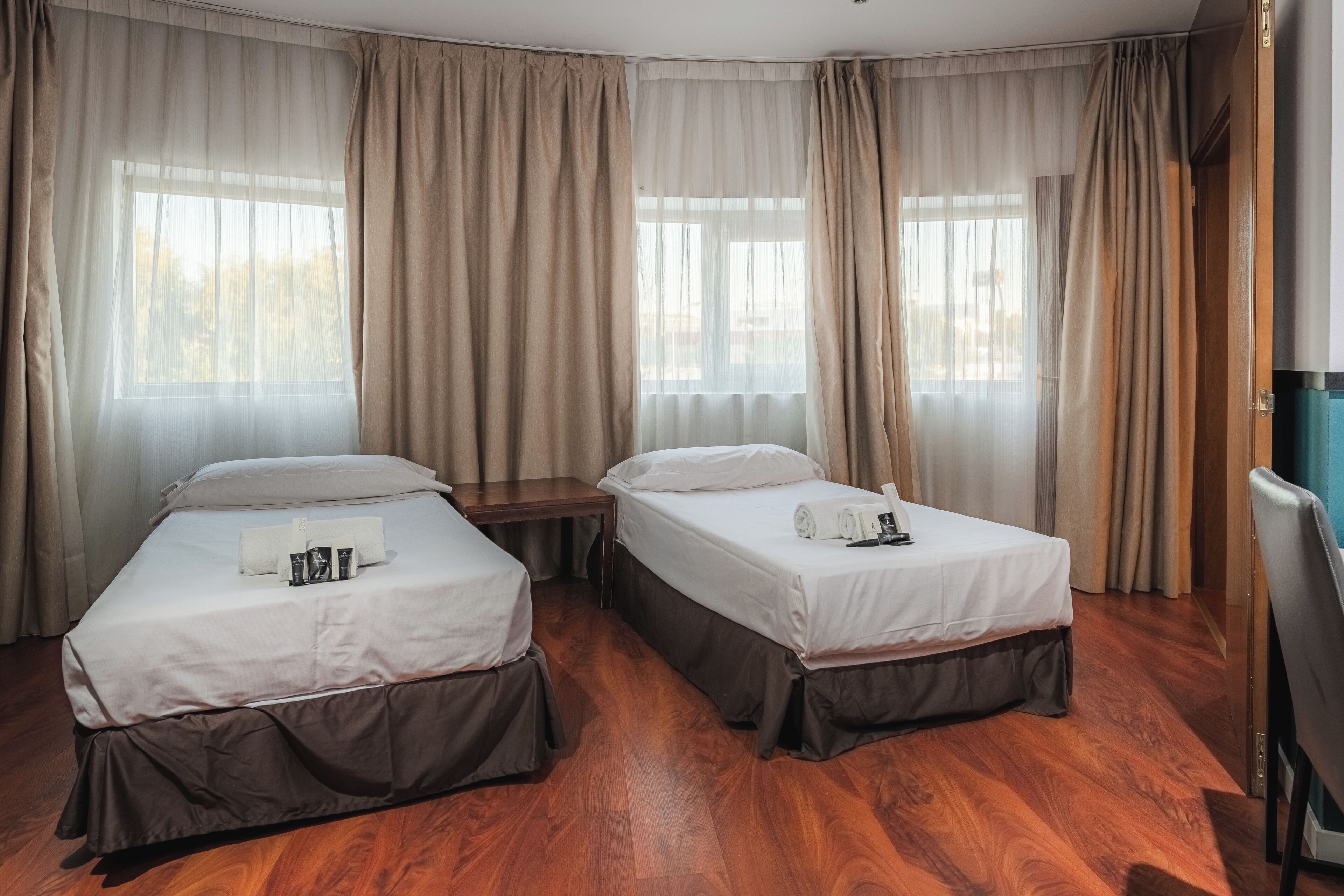 Habitación de hotel con dos camas, suelo de madera y ventanas amplias con cortinas.