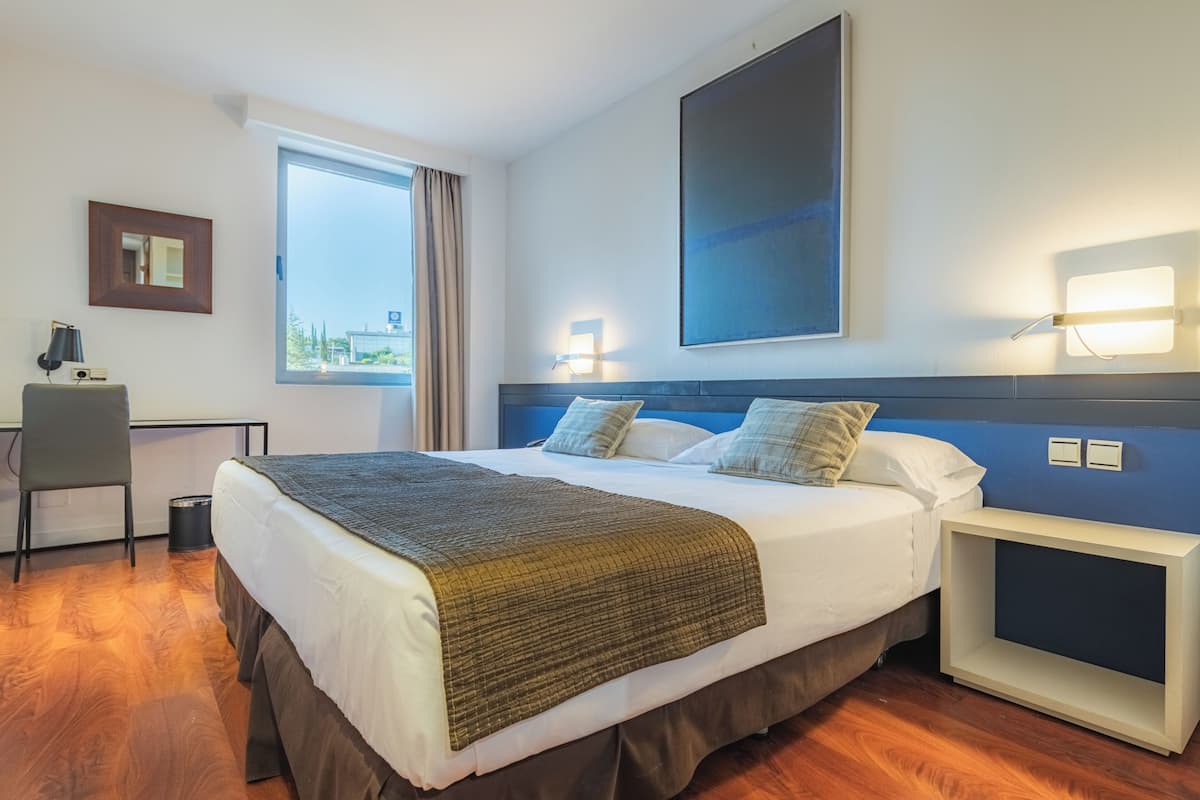Une chambre d’hôtel avec un grand lit, un bureau, une chaise, une fenêtre avec des rideaux et une œuvre d’art accrochée au mur.