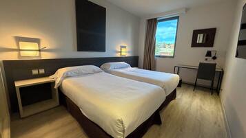 Habitación de hotel con dos camas, un escritorio, una silla, un ventanal con cortinas y luces en la pared.