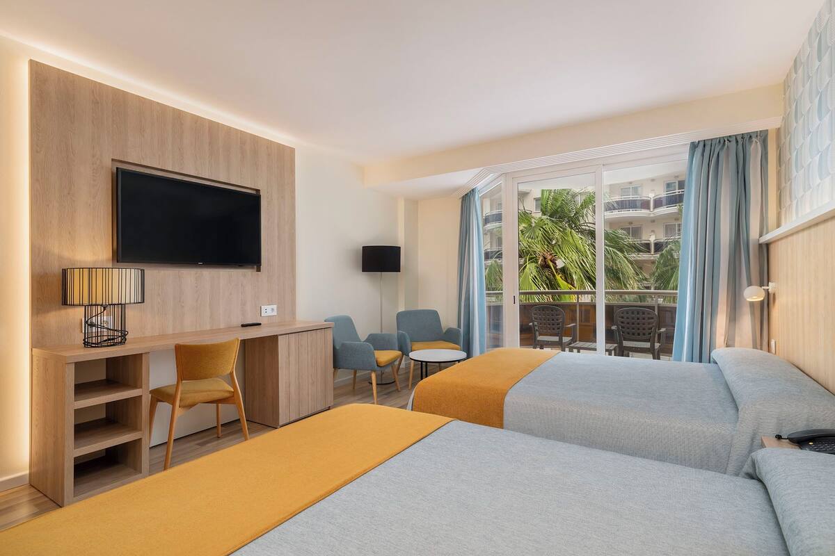 A hotel room with two beds, a desk with a TV, a chair, a small table, and a balcony with a view of the outside.