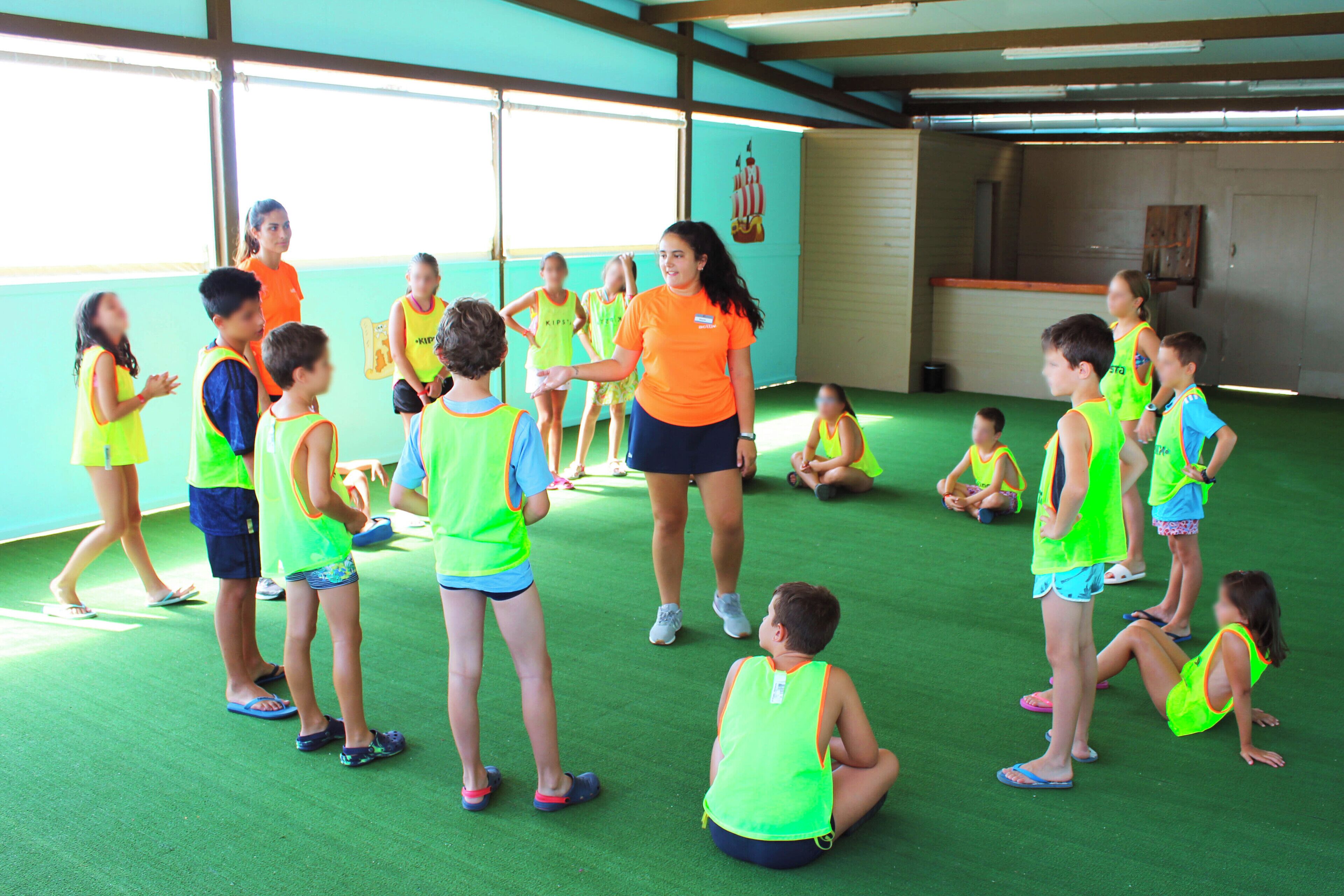 Un grupo de niños en una habitación con suelo verde y un ventanal grande, vistiendo chalecos llamativos.
