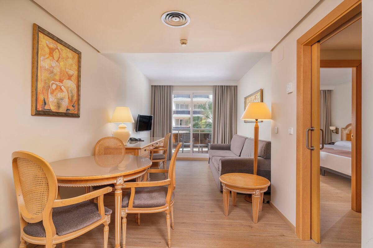 A hotel room with a dining area, a sofa, a TV, and a balcony with curtains.