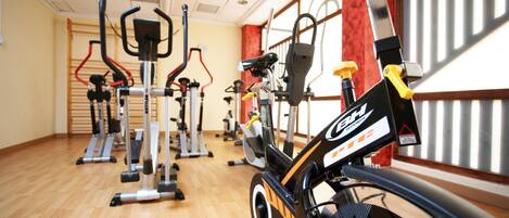 Un gimnasio con diversos equipos de ejercicio, como bicicletas estáticas y máquinas de cardio, dispuesto en un amplio espacio con suelo de madera y grandes ventanales.