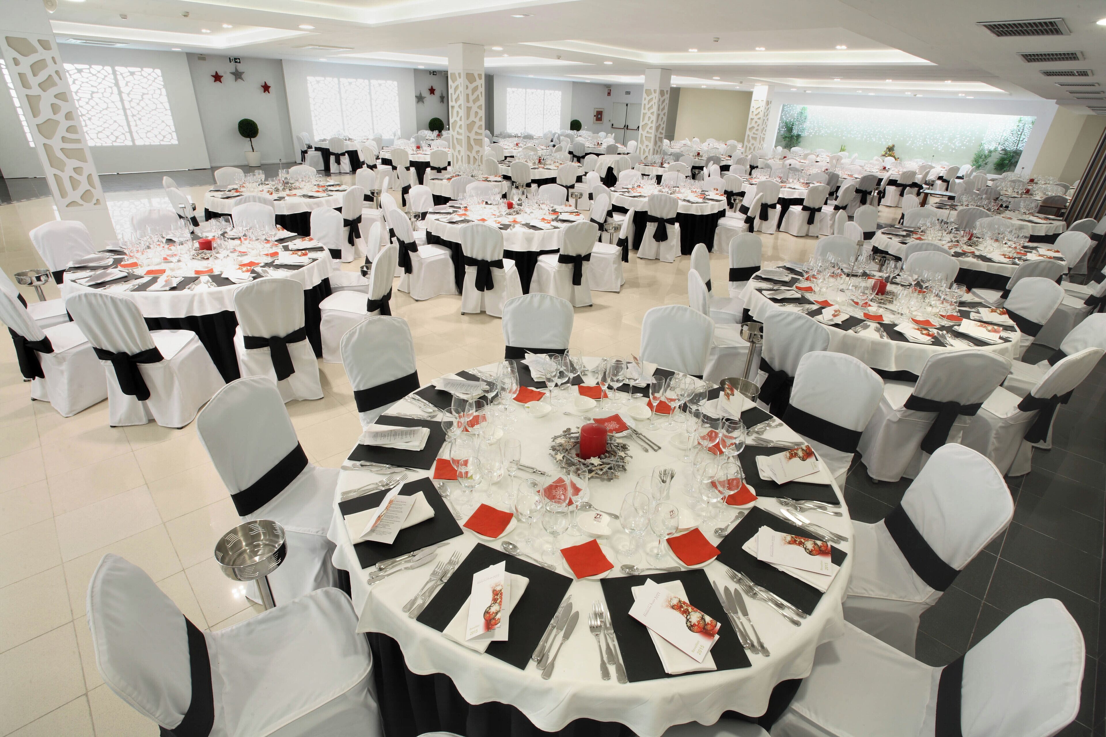 Un salón de banquetes con mesas redondas dispuestas para un evento formal, con manteles blancos, cintas negras y cubertería.