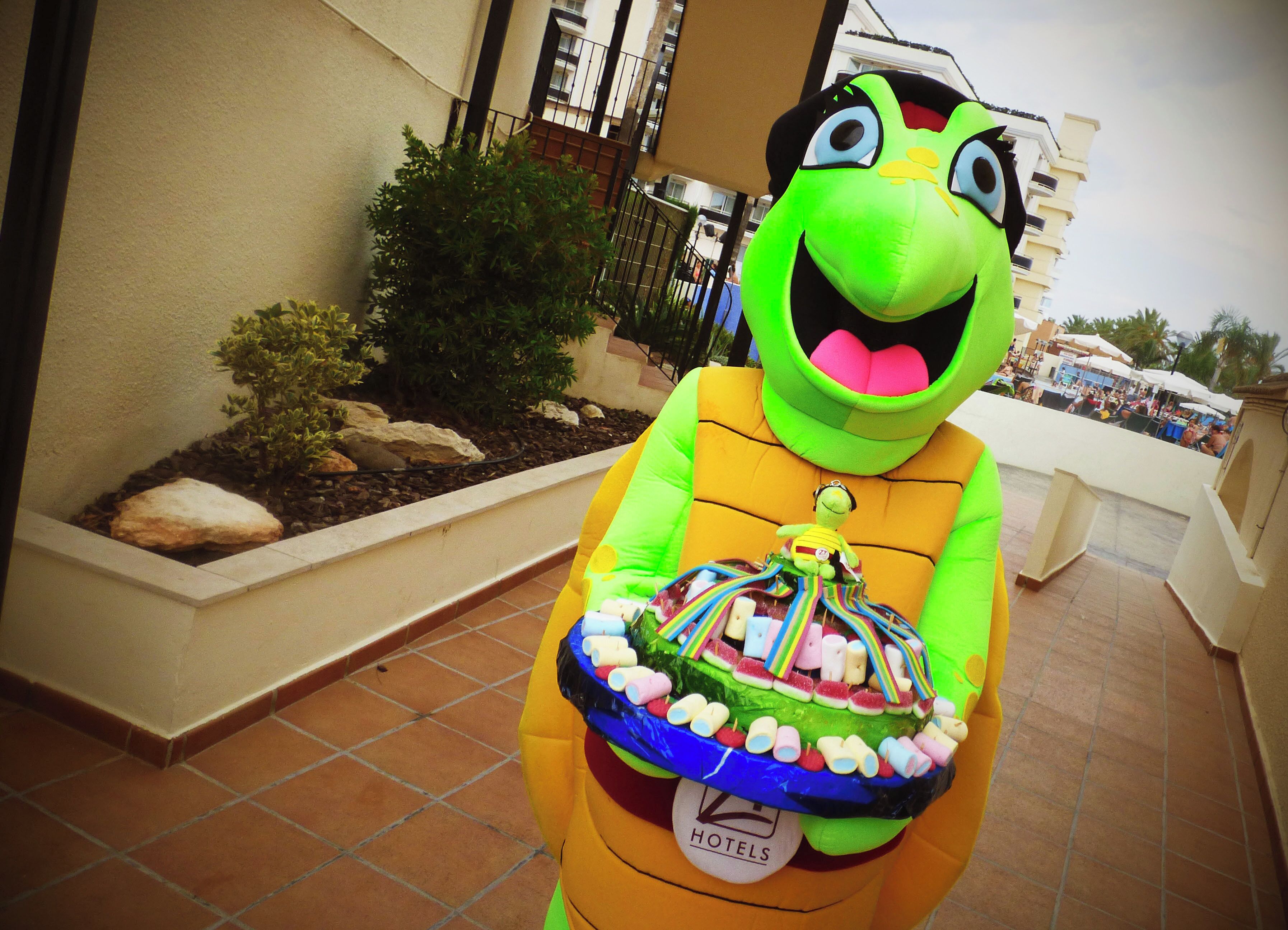 A mascot holding a colorful cake with a hotel sign on it.