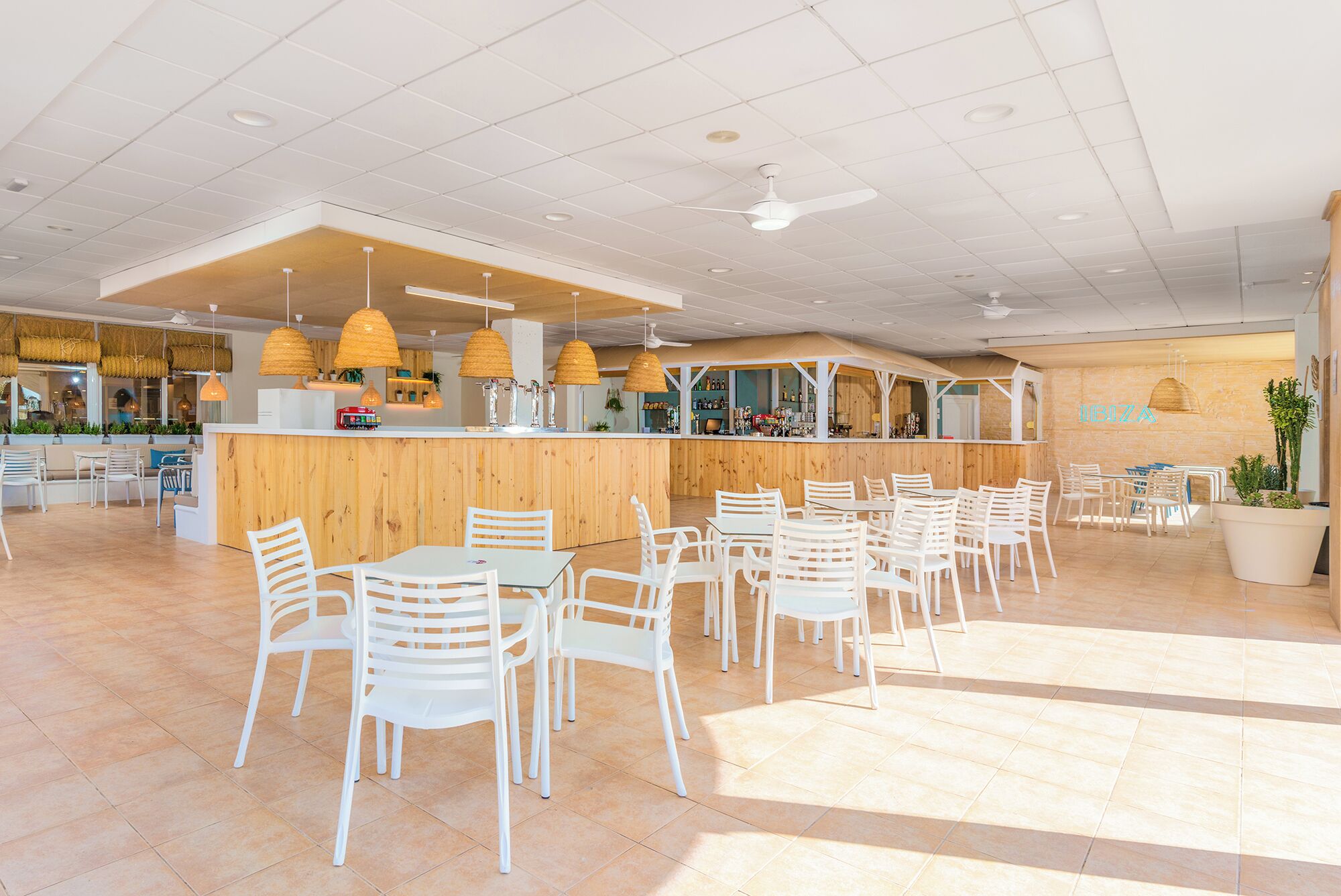 Un restaurante espacioso con una barra de madera, sillas blancas y lámparas de mimbre colgantes.