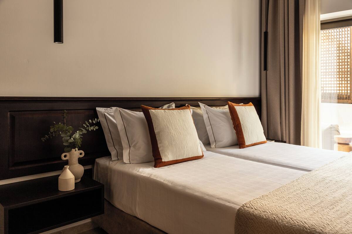 A hotel room with a double bed, a nightstand with a vase and a small plant, and a window with curtains.