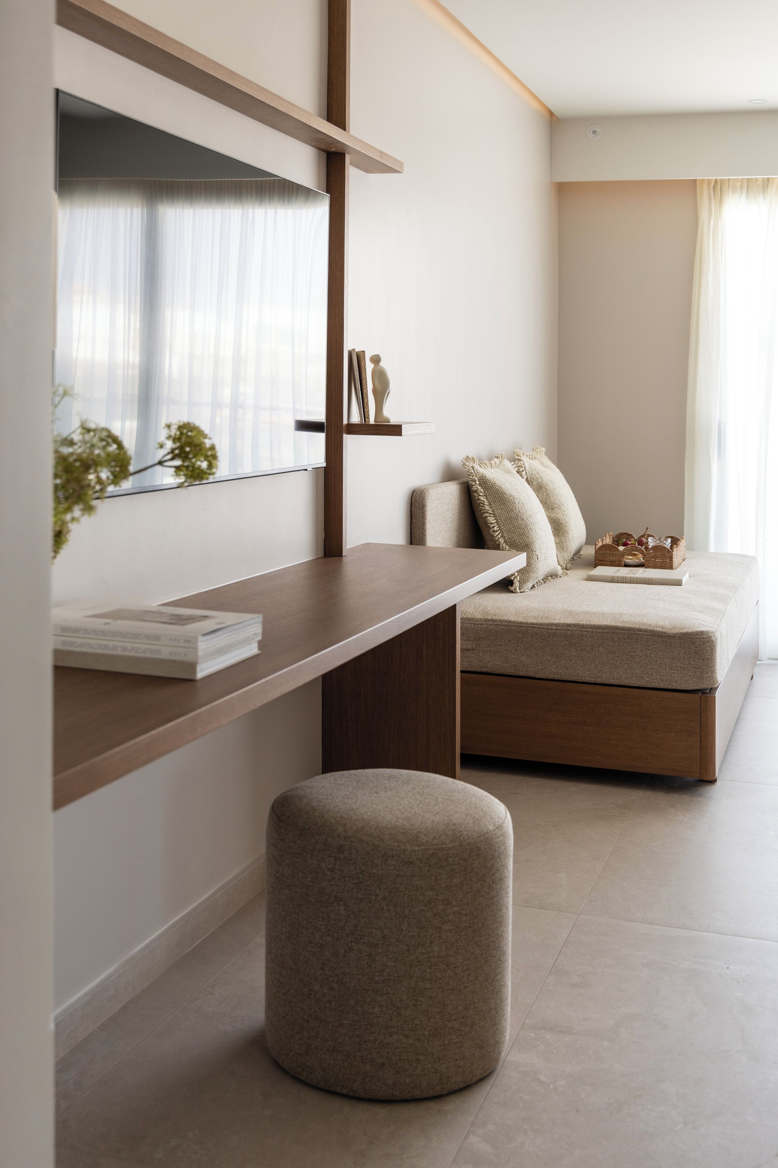 Una habitación moderna con un escritorio de madera, un banco acolchado, un pequeño reposapiés y una cama con cabecera color beige.