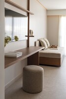 Una habitación moderna con un escritorio de madera, un banco acolchado, un pequeño reposapiés y una cama con cabecera color beige.
