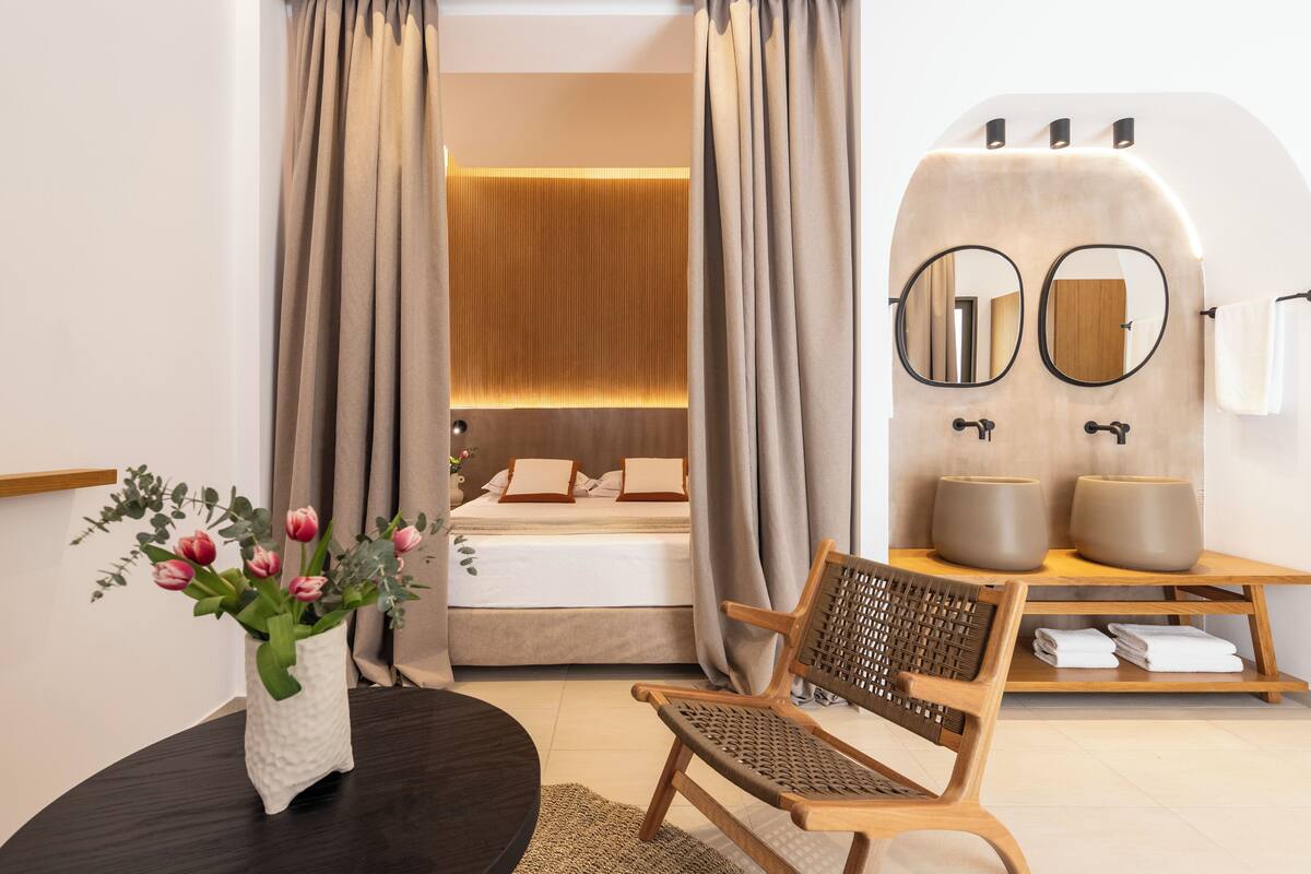 A modern hotel room with a bed, a wooden chair, a vanity with mirrors, and a vase of flowers.