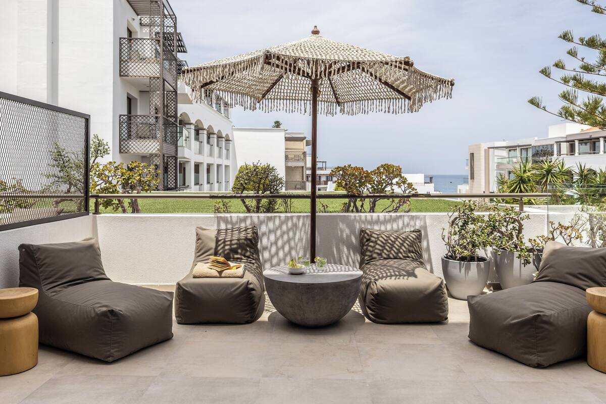 A modern outdoor seating area with a round table and two sofas, a large umbrella, and a view of the sea.