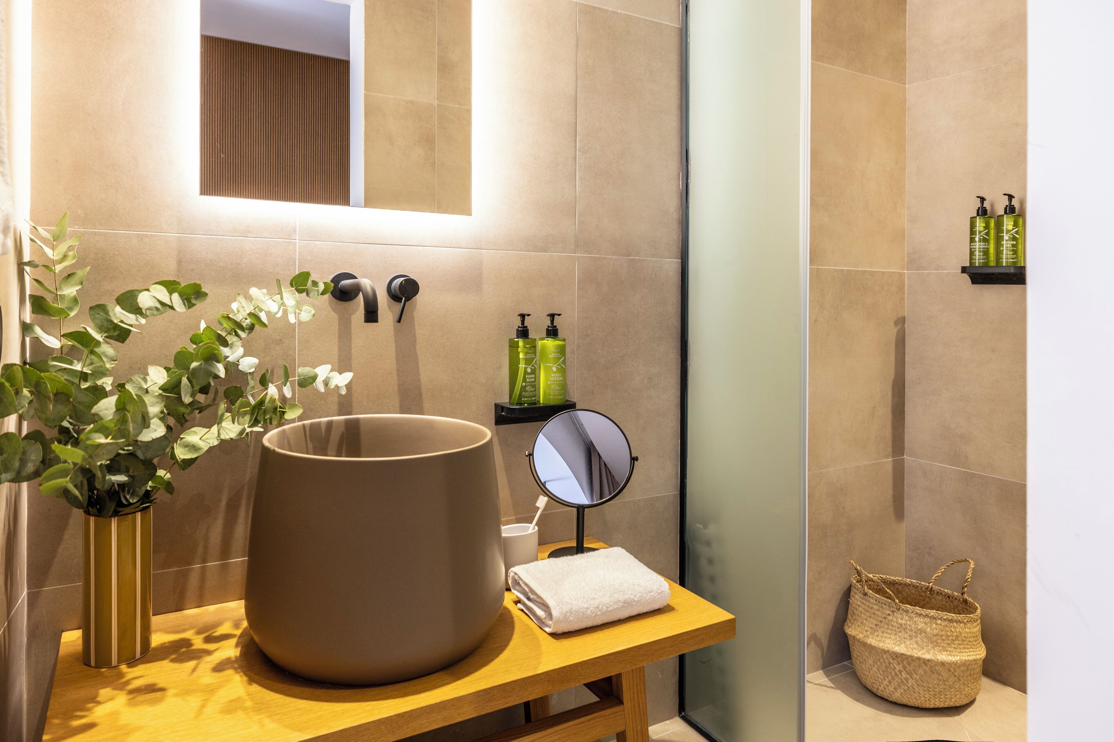 A bathroom with a large beige sink, a mirror, a small round mirror, a plant, toiletries, and a woven basket.