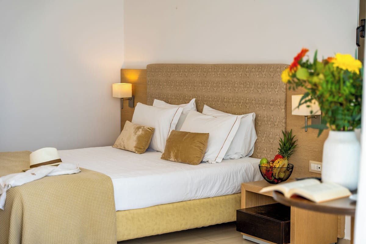 A neatly made bed with white and brown bedding, a bedside table with a fruit bowl, and a lamp on the wall.