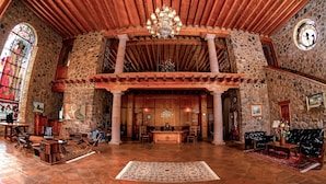Un vestíbulo espacioso con techo alto, paredes de piedra y techo de madera. Se observa un candelabro, un mostrador de recepción y un tapete grande en el suelo.